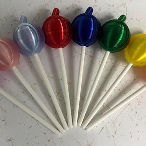 May include: A collection of nine spherical lollipops arranged in a fan shape, each featuring a distinct solid color including purple, pink, light blue, red, dark blue, green, yellow, and orange, all attached to thin white plastic sticks.