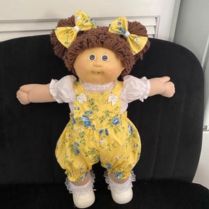 Puede incluir: Una mu&ntilde;eca con cabello de lana marr&oacute;n peinado en dos coletas con lazos florales amarillos. La mu&ntilde;eca viste una blusa blanca de manga corta con ribete de encaje bajo un peto amarillo con estampado floral azul y verde, calcetines blancos con encaje y zapatos blancos.