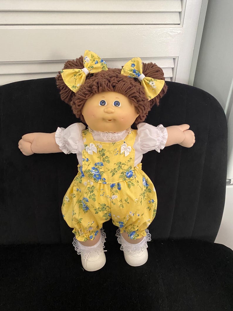 Puede incluir: Una mu&ntilde;eca con cabello de lana marr&oacute;n peinado en dos coletas con lazos florales amarillos. La mu&ntilde;eca viste una blusa blanca de manga corta con ribete de encaje bajo un peto amarillo con estampado floral azul y verde, calcetines blancos con encaje y zapatos blancos.