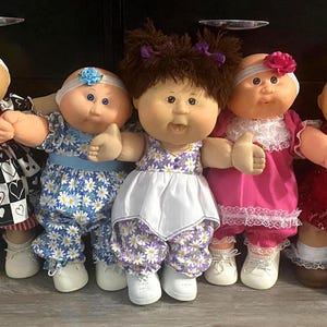 May include: Five dolls with round faces and yarn hair, each wearing a unique outfit. The dolls feature various hair colors, including brown and auburn, and are dressed in patterned dresses and headbands with white shoes and socks on their feet.