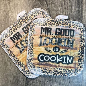 May include: Two brown and beige pot holders with a leopard print border. The pot holders have a woodgrain background with the text "Mr. Good Lookin' Is Cookin'" in black.
