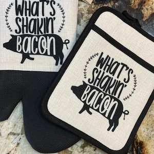 Peut inclure: Deux maniques de cuisine avec un motif de cochon noir et blanc. Les maniques portent le texte "What's Shakin' Bacon".