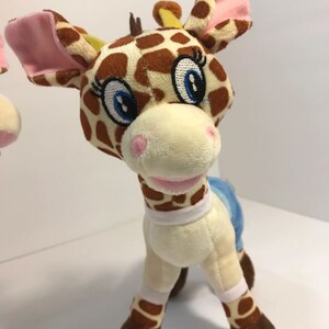 May include: A brown and yellow giraffe plush toy with pink ears, blue and white striped pants, and big blue eyes. The giraffe is standing on a white background.