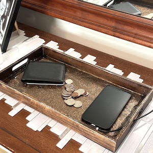 May include: A rectangular wooden tray with a dark brown finish and an ornate, embossed floral pattern on the base. Inside the tray sits a black leather wallet, a small pile of silver-colored coins, and a black smartphone connected to a charging cable.
