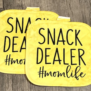 May include: Two yellow square pot holders with black text. The text on the pot holders reads "Snack Dealer #momlife".