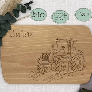 Könnte beinhalten: Ein Holzbrett zum Schneiden mit dem Namen "Julian" eingraviert. Das Brett zeigt eine Strichzeichnung eines Traktors mit den Worten "bio", "100% FSC" und "fair" in grünen Kreisen über dem Traktor.