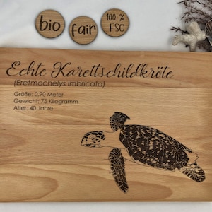 Op de afbeelding: Een houten snijplank met een lasergegraveerd beeld van een zeeschildpad. De plank is gelabeld "Echte Karettschildkröte (Eretmochelys imbricata)" en bevat informatie over de grootte, het gewicht en de leeftijd van de schildpad. De plank heeft ook drie kleine ronde houten labels met de tekst "bio", "fair" en "100% FSC".