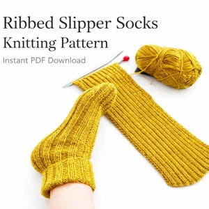 Può includere: Calzino da pantofola lavorato a maglia giallo senape con design a coste, parzialmente completato, con ferri da maglia e un gomitolo di lana. Il testo "Ribbed Slipper Socks Knitting Pattern" è in alto, con "Instant PDF Download" sotto.