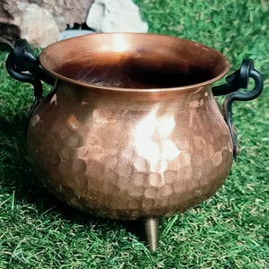 Könnte beinhalten: Ein gehämmertes Kupferkessel mit rundem Körper und drei kurzen Beinen. Der Kessel hat zwei schwarze, gebogene Griffe und steht auf grünem Gras. Das Kupfer hat einen warmen, rotbraunen Farbton.