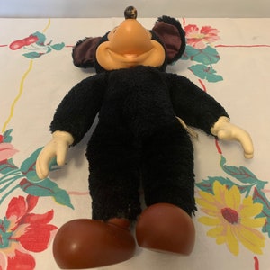 May include: A vintage plush mouse toy with a black body, large brown feet, and cream-coloured hands. The mouse has a tan face, large black ears with a maroon interior, and a small brown ball on top of its head. The toy is on a floral patterned tablecloth.