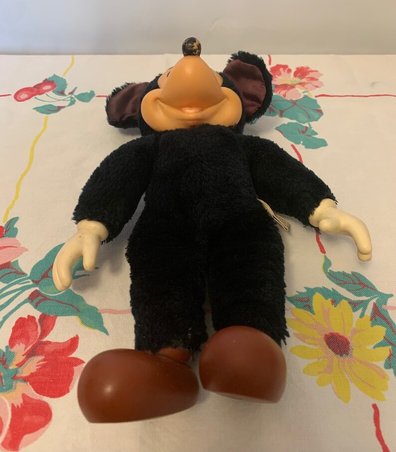 May include: A vintage plush mouse toy with a black body, large brown feet, and cream-coloured hands. The mouse has a tan face, large black ears with a maroon interior, and a small brown ball on top of its head. The toy is on a floral patterned tablecloth.
