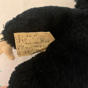 May include: A black plush toy with a tag attached. The tag has text that is difficult to read. The plush toy is dark and fluffy. The tag is rectangular and beige.