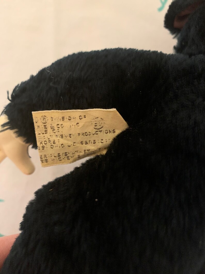 May include: A black plush toy with a tag attached. The tag has text that is difficult to read. The plush toy is dark and fluffy. The tag is rectangular and beige.