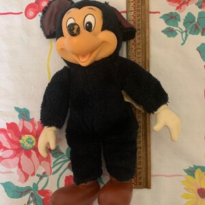 May include: Vintage Mickey Mouse plush doll with black fur, a tan face, and brown shoes. The doll has a smiling expression and cream-coloured hands. A wooden ruler is next to the doll.