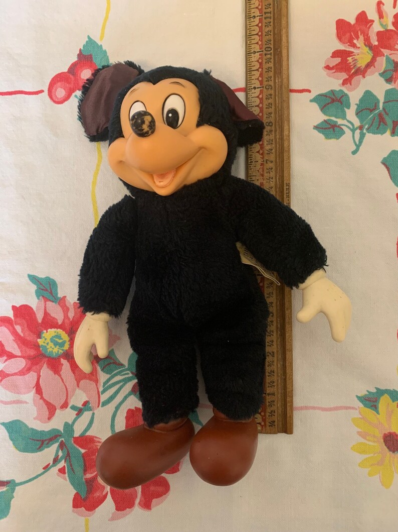 May include: Vintage Mickey Mouse plush doll with black fur, a tan face, and brown shoes. The doll has a smiling expression and cream-coloured hands. A wooden ruler is next to the doll.