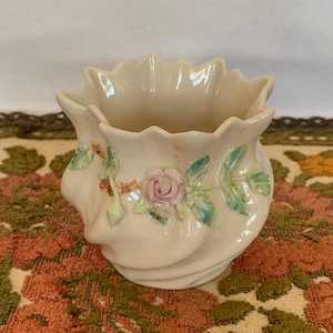 May include: Cream-colored ceramic planter with a ruffled rim and a swirling design. The planter is adorned with a pink rose and green leaves. A decorative floral accent adds a touch of elegance to this small, vintage container.