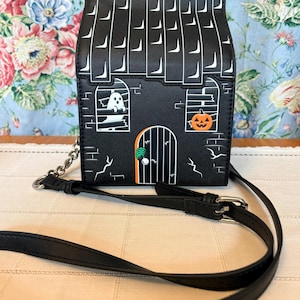 May include: A black crossbody bag shaped like a house with a roof pattern, featuring a white ghost in a window, an orange jack-o'-lantern in another window, and a green hand reaching for the door handle. It has a black adjustable shoulder strap.