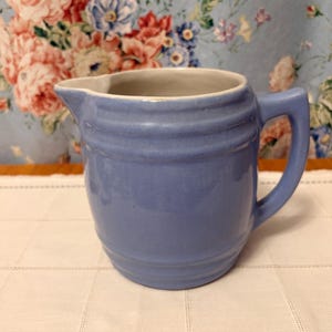 May include: A light blue ceramic pitcher with a glossy finish, featuring a rounded body with horizontal ribbed detailing, a wide pouring spout, and a sturdy side handle. The interior of the pitcher is a smooth, off-white color.