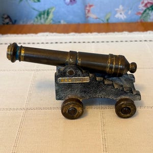 May include: A miniature bronze cannon with a dark gray carriage and four wheels. The cannon barrel has gold accents and the text "U.S.S. CONSTITUTION" is visible on the carriage. The cannon is a decorative item.