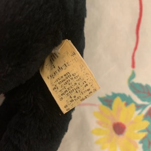 May include: A close-up of a black plush toy with a rectangular tag attached. The tag is beige with faded text. The plush toy has a soft, furry texture.
