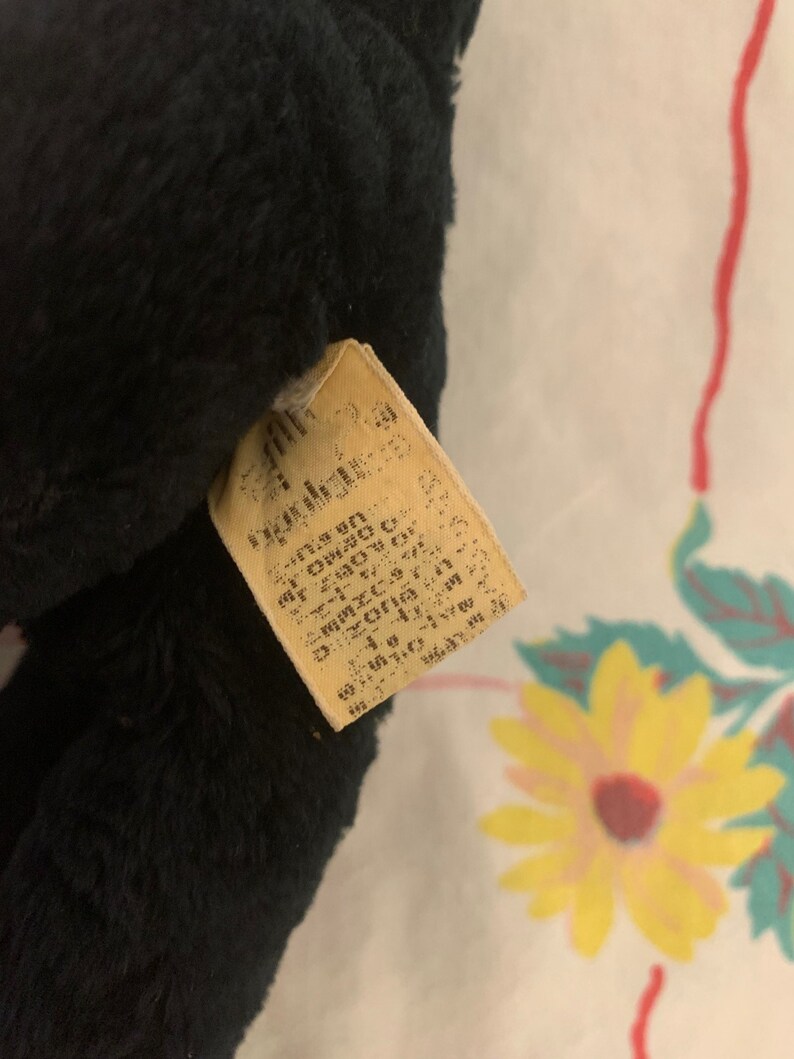 May include: A close-up of a black plush toy with a rectangular tag attached. The tag is beige with faded text. The plush toy has a soft, furry texture.