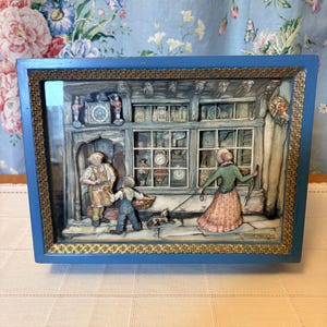 May include: A rectangular blue shadow box frame with a gold decorative border containing a three-dimensional diorama of a clockmaker shop scene. The scene features a shopkeeper, a child with a basket, and a person walking a dog past the storefront.