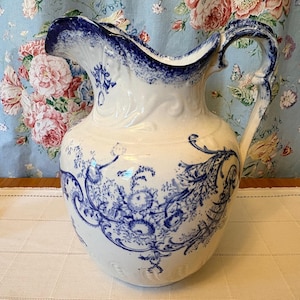 May include: A white ceramic pitcher with blue floral patterns and a blue rim. The pitcher has a large handle and a wide spout. The floral design is intricate and covers most of the pitcher's surface. The pitcher is likely vintage or antique.