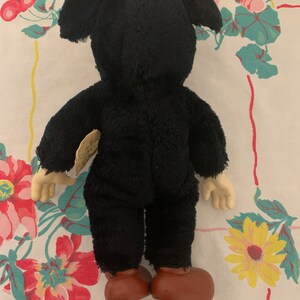May include: A vintage black plush toy with large ears, tan hands, and brown shoes. The toy is viewed from the back, revealing its plush texture and design. A small tag is attached to the toy.