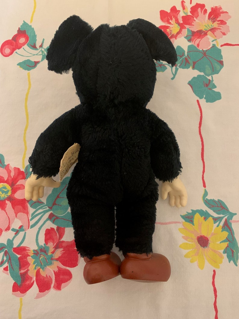 May include: A vintage black plush toy with large ears, tan hands, and brown shoes. The toy is viewed from the back, revealing its plush texture and design. A small tag is attached to the toy.