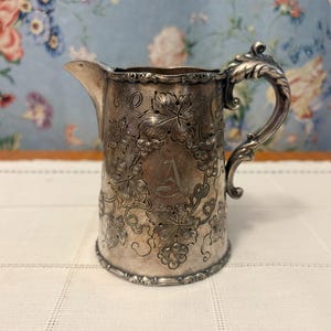 May include: A silver-toned metal creamer pitcher featuring an embossed pattern of grape leaves and vines. The pitcher has a curved handle, a pointed spout, and a central engraved letter A on the front surface with a tarnished finish.