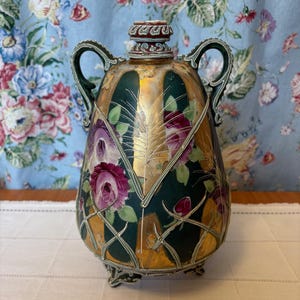 May include: An antique decorative vase with a dark green and gold finish, featuring hand-painted purple roses and a leaf design. The vase has two handles and rests on small feet, showcasing an ornate, vintage aesthetic.