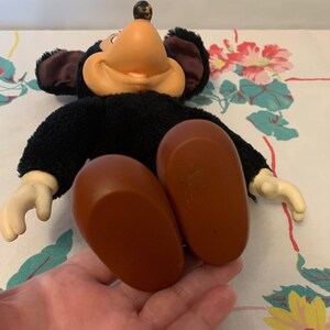 May include: Vintage Mickey Mouse plush doll with black fur, a tan face, and large brown feet. The doll has white gloves and large black ears. The doll is lying on a floral patterned tablecloth.