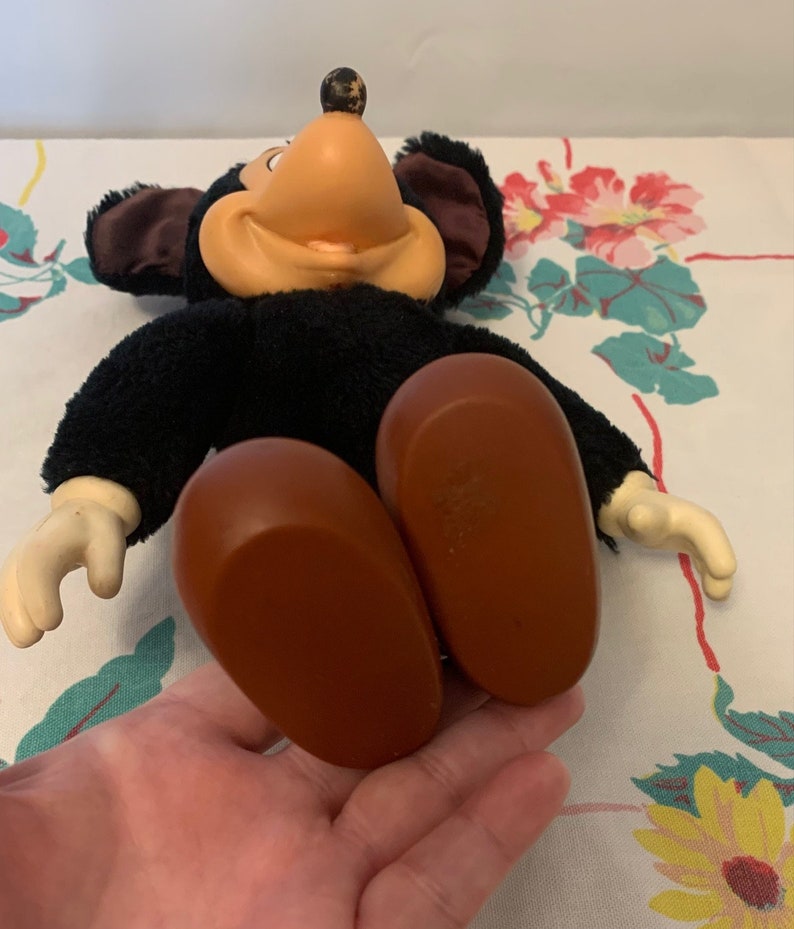 May include: Vintage Mickey Mouse plush doll with black fur, a tan face, and large brown feet. The doll has white gloves and large black ears. The doll is lying on a floral patterned tablecloth.