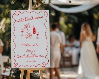 May include: A white sign with red lettering that says "That's Amore!" and features a drawing of a pizza, wine glasses, and a bottle of wine. The sign also says "Welcome to Lucy's Bridal Shower 10.25.2024".