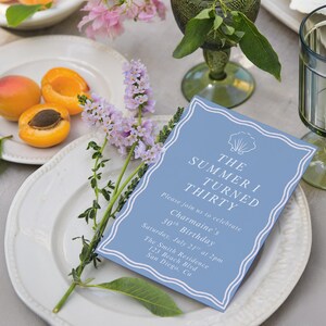 May include: A light blue invitation card with white text that reads "The Summer I Turned Thirty". The card is on a white plate with apricots and purple flowers. A green glass and a second plate are also visible.