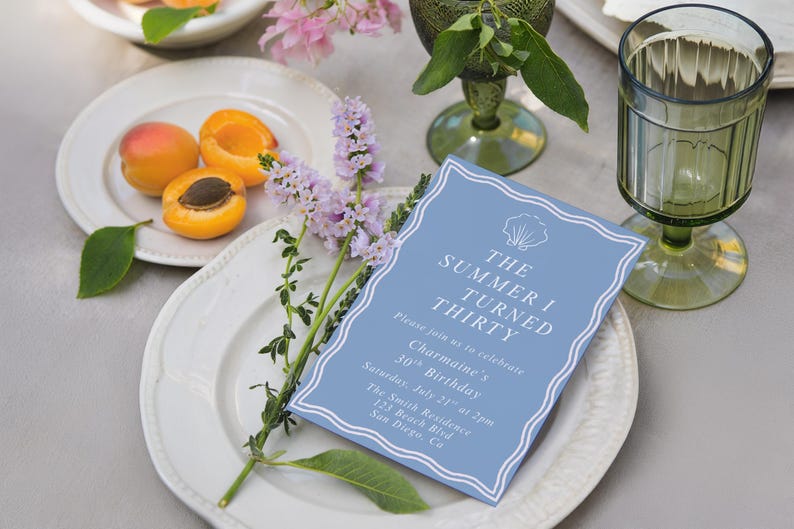 May include: A light blue invitation card with white text that reads "The Summer I Turned Thirty". The card is on a white plate with apricots and purple flowers. A green glass and a second plate are also visible.
