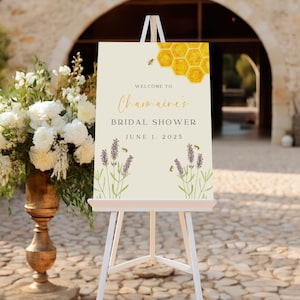 Może przedstawiać: Kremowy znak powitalny na wieczór panieński, umieszczony na białym sztaludze. Znak zawiera tekst "Welcome to Charmaine's Bridal Shower June 1, 2025." Zawiera ilustracje plastra miodu, pszczół i lawendy.