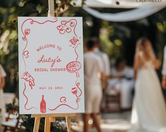 May include: A white sign with red lettering on an easel announcing a bridal shower for Lucy. The sign features illustrations of pizza, wine glasses, and tomatoes.