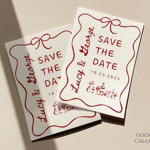 Può includere: Due biglietti bianchi "Save the Date" con testo rosso e un fiocco rosso. Il testo recita "Lucy & George Save the Date 10.25.2025". I biglietti hanno un bordo ondulato e una piccola illustrazione di bicchieri di vino e bottiglie.