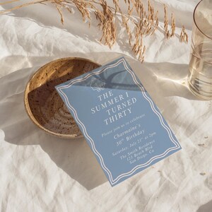 May include: A light blue invitation card with white decorative borders and text that reads "THE SUMMER I TURNED THIRTY". The card is for a 30th birthday celebration. A ceramic bowl and a glass are also in the image.