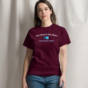 May include: A maroon t-shirt with the text "Tej Chaws Dej Xiav, Land of Blue Water" in white and light blue. The shirt is paired with blue jeans and white sneakers. The person is standing in front of a white curtain.