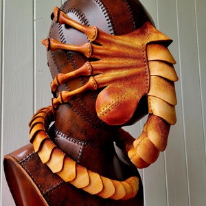 May include: A brown leather mask with a detailed design of a creature with multiple arms and a segmented tail. The mask is displayed on a brown leather mannequin.