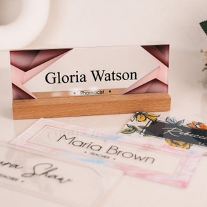 May include: A clear acrylic desk nameplate with a pink and brown geometric design. The nameplate has a wooden base and the text "Gloria Watson, Pharmacist" is printed on the front.