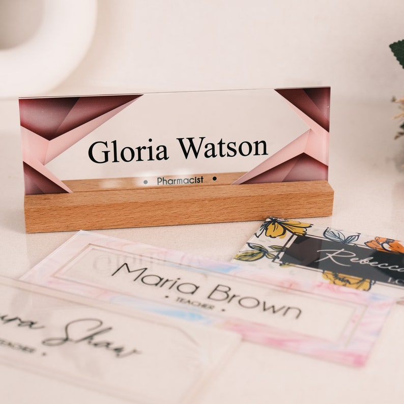 May include: A clear acrylic desk nameplate with a pink and brown geometric design. The nameplate has a wooden base and the text "Gloria Watson, Pharmacist" is printed on the front.