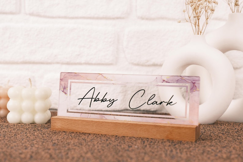 May include: A clear acrylic nameplate with a pink and purple marble design. The nameplate has a wooden base and the name "Abby Clark" is written in black cursive.