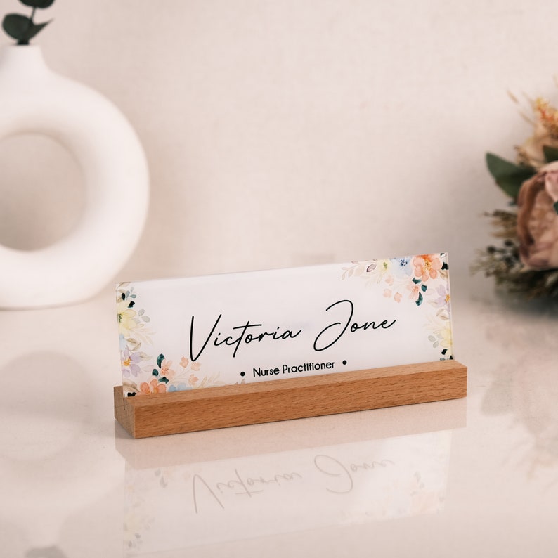 May include: A white acrylic desk nameplate with a floral design and the text "Victoria Jones - Nurse Practitioner" on a wooden stand.
