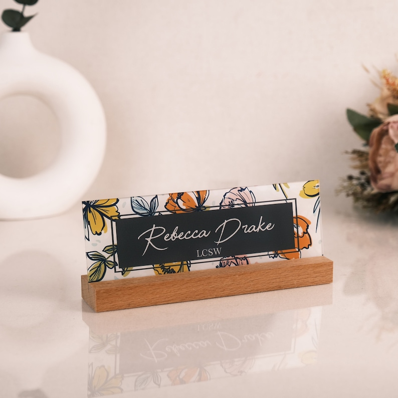 May include: A wooden desk nameplate with a floral design and the name "Rebecca Drake, LCSW" printed on a black background.