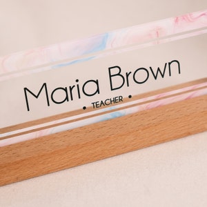 May include: A clear acrylic desk nameplate with a pink and blue marbled design. The nameplate has a wooden base and the text "Maria Brown - TEACHER" is printed on the acrylic.