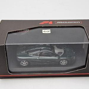 Op de afbeelding: Een donkergroene McLaren F1 modelauto in een zwarte displaydoos met een doorzichtig plastic venster. De doos heeft rode en witte tekst met de tekst "F1 McLaren". De modelauto heeft zilveren wielen.