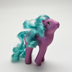 May include: A purple toy pony with teal blue hair and a matching tail. The pony has a small, decorative design on its cheek and a pink eye. The toy is standing on a white surface.